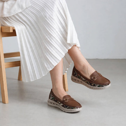 Woman sitting on wooden chair wearing brown woven loafers with white pleated skirt